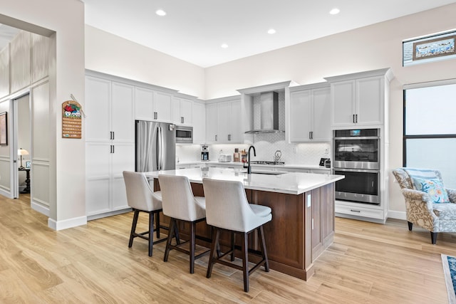 kitchen with a breakfast bar, wall chimney range hood, appliances with stainless steel finishes, backsplash, and a center island with sink