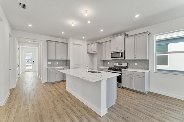 kitchen with gray cabinets, appliances with stainless steel finishes, sink, a kitchen island with sink, and light stone counters