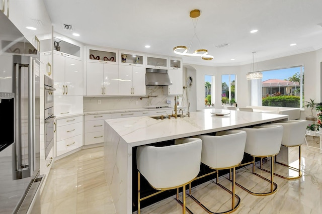 kitchen featuring white cabinetry, tasteful backsplash, light stone counters, decorative light fixtures, and a center island with sink