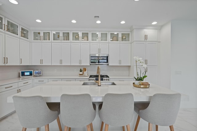 kitchen featuring a kitchen island with sink, stainless steel microwave, glass insert cabinets, and white cabinets