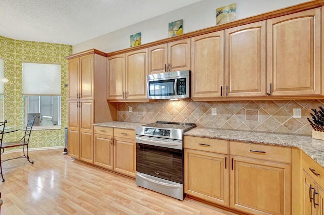 kitchen with wallpapered walls, baseboards, appliances with stainless steel finishes, light stone counters, and light wood-type flooring