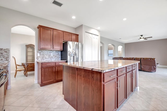 kitchen featuring light tile patterned floors, backsplash, stainless steel fridge, an island with sink, and light stone countertops