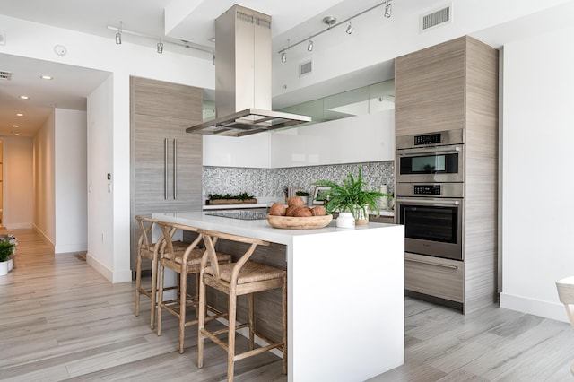 kitchen featuring tasteful backsplash, a breakfast bar area, double oven, island exhaust hood, and light wood-type flooring