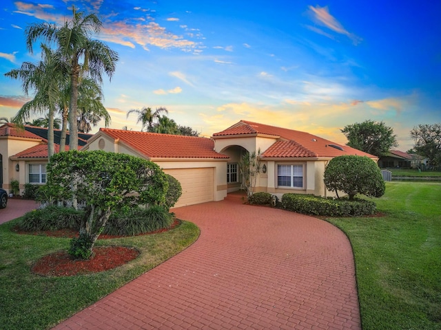 mediterranean / spanish home with decorative driveway, stucco siding, a lawn, an attached garage, and a tiled roof