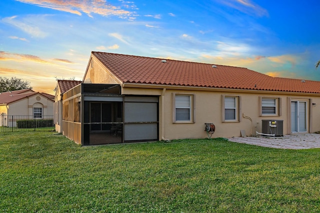 back of house at dusk featuring a lawn, a sunroom, fence, central AC, and stucco siding