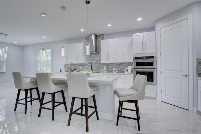 kitchen featuring hanging light fixtures, wall chimney exhaust hood, a center island with sink, and white cabinetry