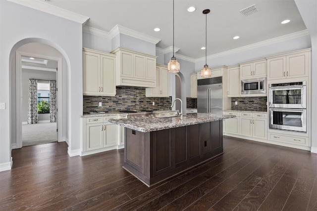 kitchen with built in appliances, arched walkways, light stone counters, cream cabinets, and a center island with sink