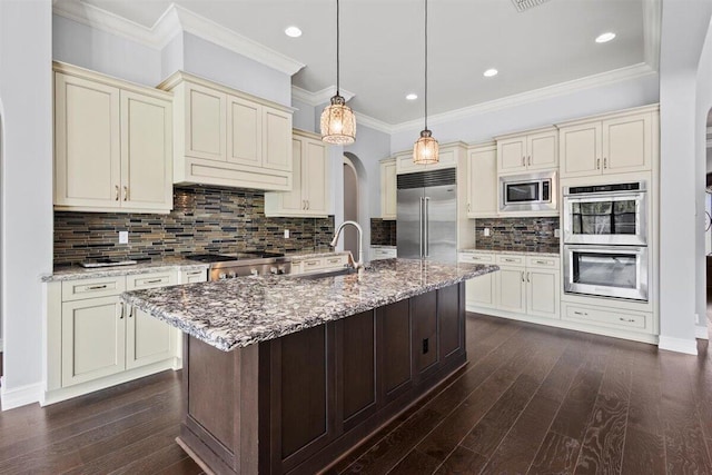 kitchen with arched walkways, dark wood finished floors, cream cabinets, light stone countertops, and built in appliances