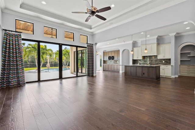 unfurnished living room featuring dark wood-type flooring, arched walkways, a raised ceiling, and a ceiling fan