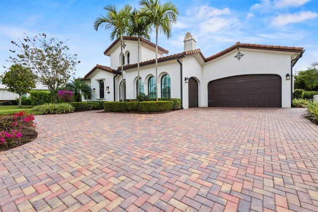 mediterranean / spanish home with an attached garage, a tile roof, decorative driveway, stucco siding, and a chimney