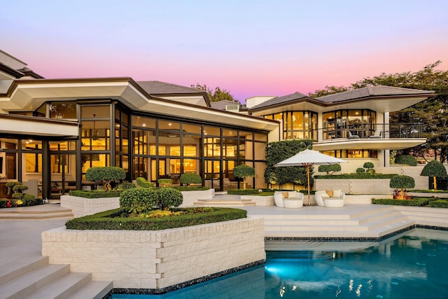 back of house at dusk featuring a patio area, an outdoor pool, and a balcony