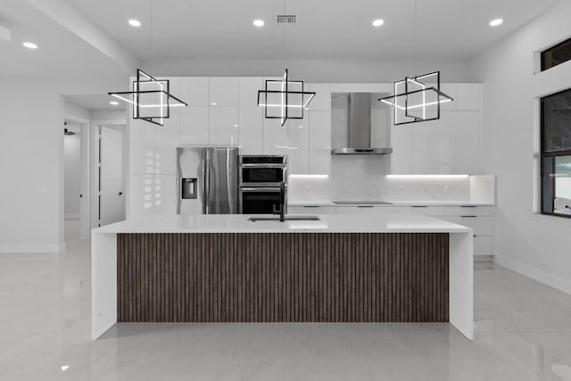 kitchen with white cabinetry, an island with sink, hanging light fixtures, appliances with stainless steel finishes, and wall chimney range hood