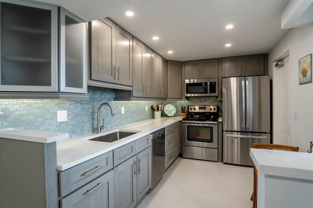 kitchen with gray cabinetry, a sink, appliances with stainless steel finishes, backsplash, and glass insert cabinets