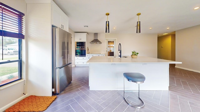 kitchen featuring appliances with stainless steel finishes, a center island with sink, wall chimney range hood, white cabinets, and pendant lighting