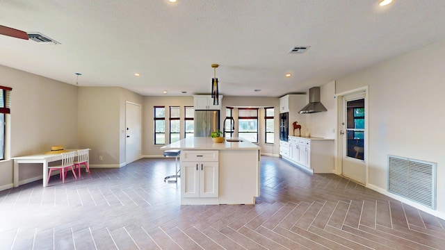 kitchen with a kitchen island with sink, hanging light fixtures, white cabinets, a breakfast bar area, and wall chimney range hood