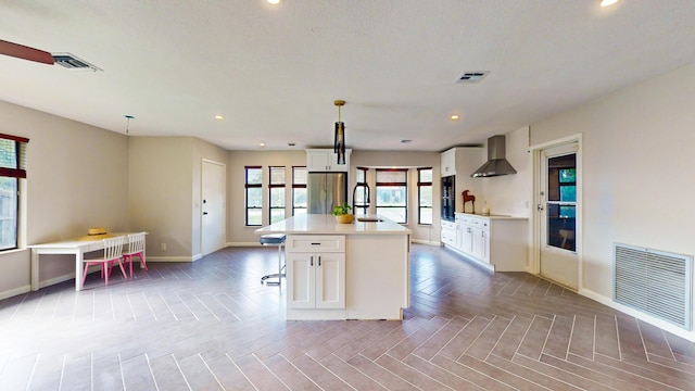 kitchen featuring wall chimney exhaust hood, black appliances, white cabinets, decorative light fixtures, and a kitchen island with sink