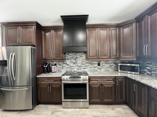 kitchen with dark brown cabinetry, tasteful backsplash, custom range hood, light stone countertops, and stainless steel appliances