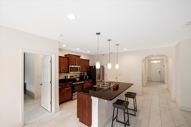 kitchen featuring arched walkways, dark countertops, appliances with stainless steel finishes, a sink, and an island with sink