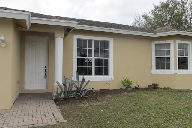 view of exterior entry with a yard, roof with shingles, and stucco siding