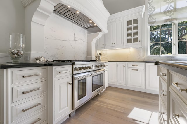 kitchen featuring light wood finished floors, dark countertops, glass insert cabinets, custom exhaust hood, and white cabinetry