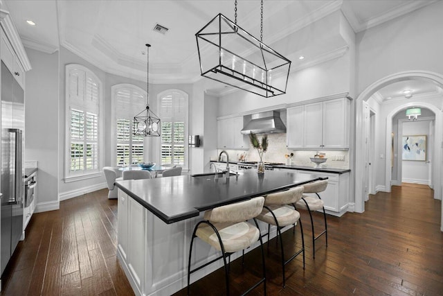 kitchen with arched walkways, white cabinets, dark countertops, wall chimney exhaust hood, and a sink