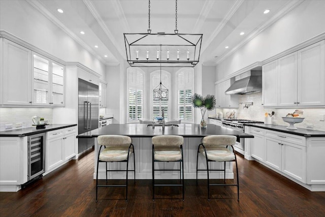 kitchen featuring wine cooler, high quality appliances, wall chimney exhaust hood, an island with sink, and dark countertops