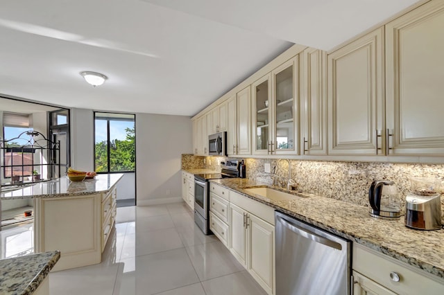 kitchen with light stone counters, glass insert cabinets, stainless steel appliances, cream cabinetry, and a sink