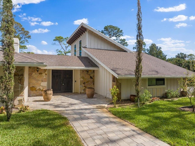 view of front of home with a shingled roof, stone siding, board and batten siding, a chimney, and a front yard