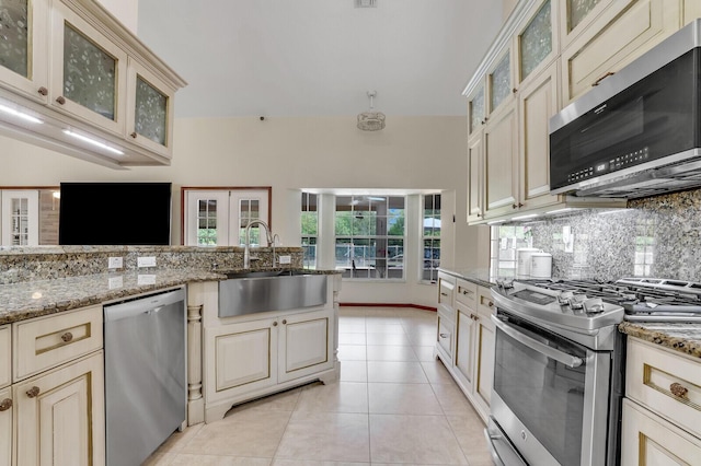 kitchen with cream cabinetry, appliances with stainless steel finishes, a sink, and glass insert cabinets