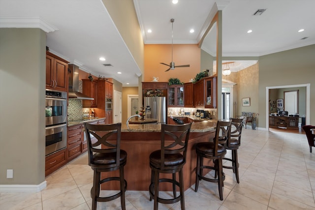 kitchen with visible vents, appliances with stainless steel finishes, glass insert cabinets, a kitchen island with sink, and wall chimney range hood