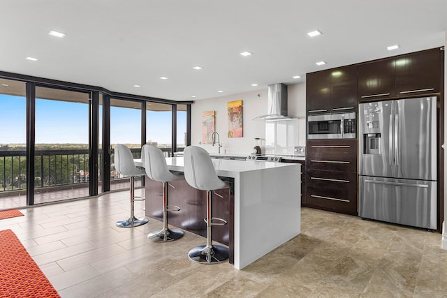 kitchen featuring a center island with sink, wall chimney exhaust hood, appliances with stainless steel finishes, expansive windows, and light countertops