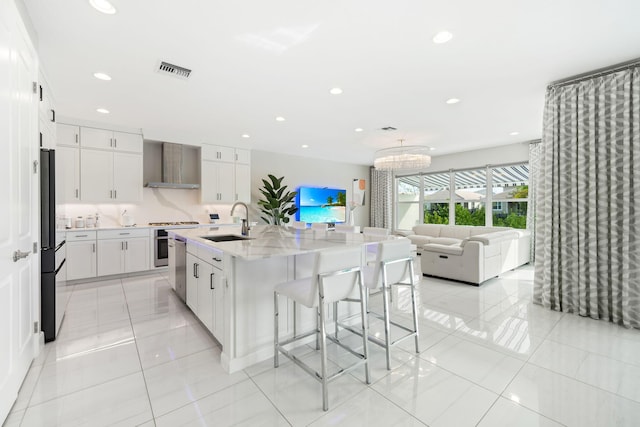 kitchen featuring visible vents, white cabinets, a sink, an island with sink, and wall chimney exhaust hood