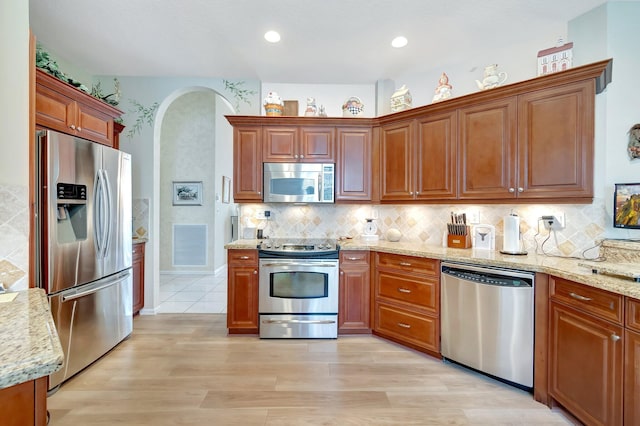 kitchen featuring tasteful backsplash, arched walkways, brown cabinetry, appliances with stainless steel finishes, and light stone countertops