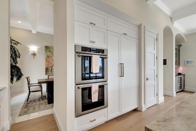 kitchen with arched walkways, double oven, ornamental molding, white cabinetry, and light wood-type flooring