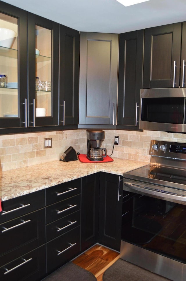kitchen with glass insert cabinets, appliances with stainless steel finishes, decorative backsplash, and dark cabinets