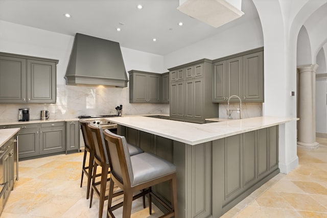 kitchen featuring arched walkways, wall chimney exhaust hood, gray cabinetry, stainless steel stove, and ornate columns