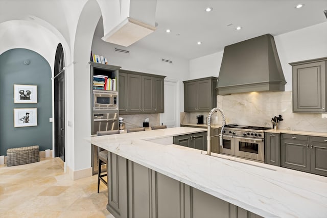 kitchen with appliances with stainless steel finishes, light stone countertops, gray cabinetry, premium range hood, and backsplash