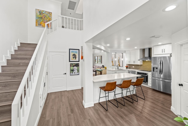 kitchen featuring wood finished floors, a peninsula, stainless steel appliances, wall chimney range hood, and a kitchen bar