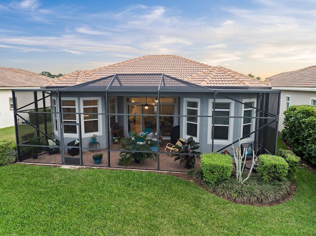 back of house at dusk with a tile roof, glass enclosure, a lawn, and a patio