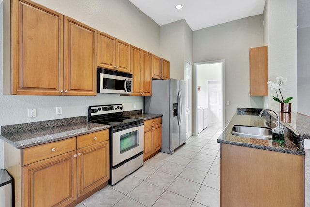 kitchen featuring light tile patterned floors, stainless steel appliances, brown cabinetry, a sink, and dark stone countertops