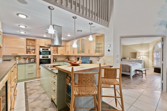 kitchen featuring glass insert cabinets, island exhaust hood, stainless steel double oven, light brown cabinetry, and green cabinets