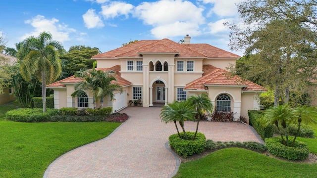 mediterranean / spanish-style house with a tile roof, decorative driveway, stucco siding, a chimney, and a front yard