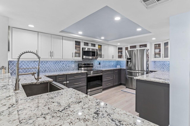 kitchen with white cabinets, appliances with stainless steel finishes, glass insert cabinets, light stone countertops, and a tray ceiling