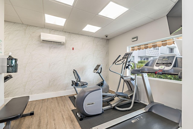 exercise room featuring a wall unit AC, a paneled ceiling, wood finished floors, baseboards, and wallpapered walls