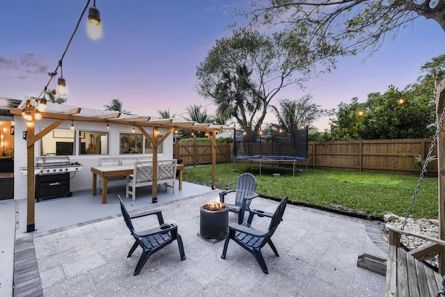 patio terrace at dusk featuring a trampoline, a lawn, a pergola, a fenced backyard, and a fire pit