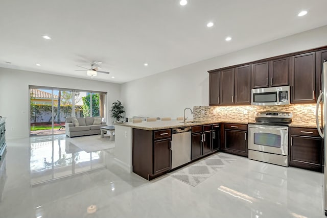 kitchen featuring dark brown cabinetry, stainless steel appliances, a sink, backsplash, and light stone countertops