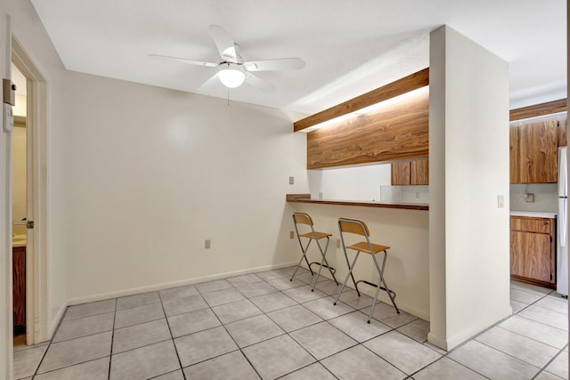 kitchen featuring light tile patterned floors, a ceiling fan, a kitchen breakfast bar, freestanding refrigerator, and brown cabinets