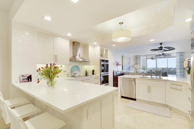 kitchen with a raised ceiling, light countertops, appliances with stainless steel finishes, wall chimney range hood, and a peninsula