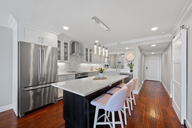 kitchen featuring wall chimney exhaust hood, glass insert cabinets, appliances with stainless steel finishes, a center island, and light countertops