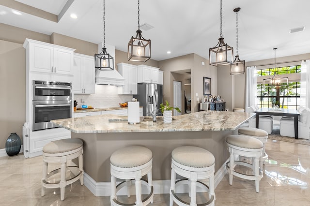 kitchen featuring light stone counters, white cabinetry, hanging light fixtures, appliances with stainless steel finishes, and a large island with sink